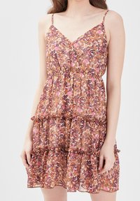 Femme portant une robe sans manches à fleurs aux teintes roses, orange et marron, avec des volants et une taille cintrée.
