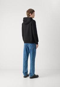 Svart hettegenser kombinert med lyseblå, løstsittende jeans og svarte sko, sett fra bak. Myk stoff med et enkelt design.