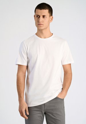 Jonge man met kort donker haar draagt een effen wit t-shirt en grijze broek, staat met één hand in de zak tegen een lichte achtergrond.