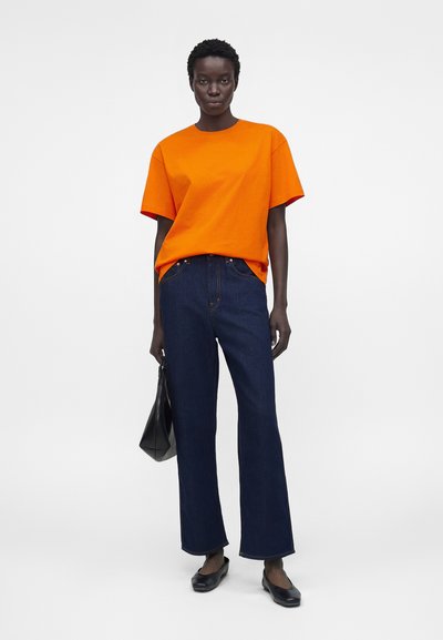 Άτομο που φοράει φωτεινή πορτοκαλί κοντομάνικη μπλούζα, σκούρα μπλε παντελόνια με φαρδιά μπάσκα, μαύρα μπαλαρίνες και κρατά μια μαύρη τσάντα.