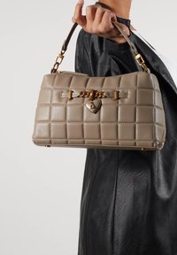 Beżowa pikowana torebka na ramię z złotym serduszkiem, o wyraźnej formie i krótkim uchwycie, znajdująca się na tle czarnej skórzanej kurtki.