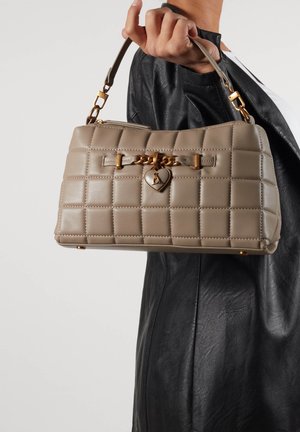 Beige gewatteerde handtas met een gouden hartbedel, met een gestructureerde vorm en korte handgreep, tegen een zwarte leren jas.