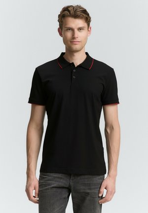 Zwarte polo met rode kraagafwerking, korte mouwen en een drieknopenlijst. Model draagt het met grijze jeans, op een effen achtergrond.