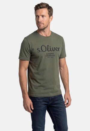 Mann trägt ein olivgrünes Kurzarm-T-Shirt mit der Aufschrift "s.Oliver Elevated Essentials Since 1969" und dunkelblaue Jeans, schaut nach links.