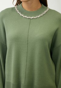 Groene gebreide trui met een hoge hals, versierd met een decoratieve kristallen kraag. De stof is zacht met een gladde textuur.