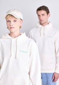 Sweat-shirts crème avec cordons de serrage et poches avant, arborant un petit logo "tommy jeans" turquoise. Une casquette beige est portée par le modèle à l'avant.