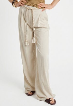 Beige Hosen mit weitem Bein, geflochtenem Seilgürtel und Quasten, kombiniert mit braunen Sandalen mit offenen Zehen und einem beigefarbenen Oberteil, Hände in der Hüfte.