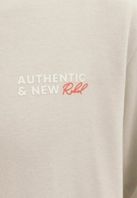 Υφασμα εκρού με το κείμενο "AUTHENTIC & NEW" σε λευκό και το "Rebel" σε κόκκινη πλάγια γραφή.