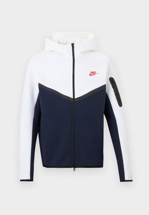 Nike-jas met capuchon, witte bovenkant, donkerblauwe onderkant, zwarte rits in het midden en een klein rood Nike-logo op de linkerborst.