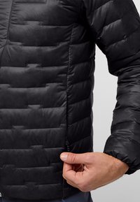 Veste noire rembourrée avec un design matelassé, dotée d'une poche zippée sur le côté. La texture est lisse avec un léger éclat, et la coupe est ajustée.