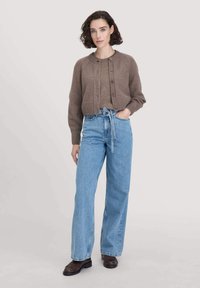 Ruskea lyhyt neulejakku, jossa on ribbireuna ja napit, päällä vaaleanruskea toppi, yhdistettynä korkeavyötäröisiin vaaleansinisiin farkkuihin ja mustiin kenkiin.