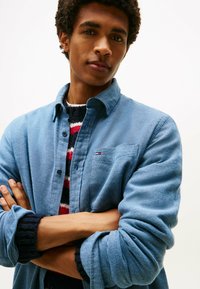 Camicia azzurra con bottoni in tessuto texturizzato, dotata di taschino e piccolo logo; indossata sopra un maglione a righe con accenti scuri.