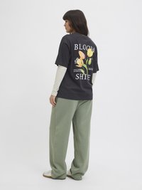 Schwarzes Baumwoll-T-Shirt mit bunten Blumenmotiven und weißem Text auf der Rückseite, kombiniert mit lockeren grünen Sweatpants und weißen Sneakern.