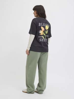 Svart bomull t-skjorte med fargerike blomster grafikk og hvit tekst på baksiden, kombinert med løse grønne joggebukser og hvite sneakers.