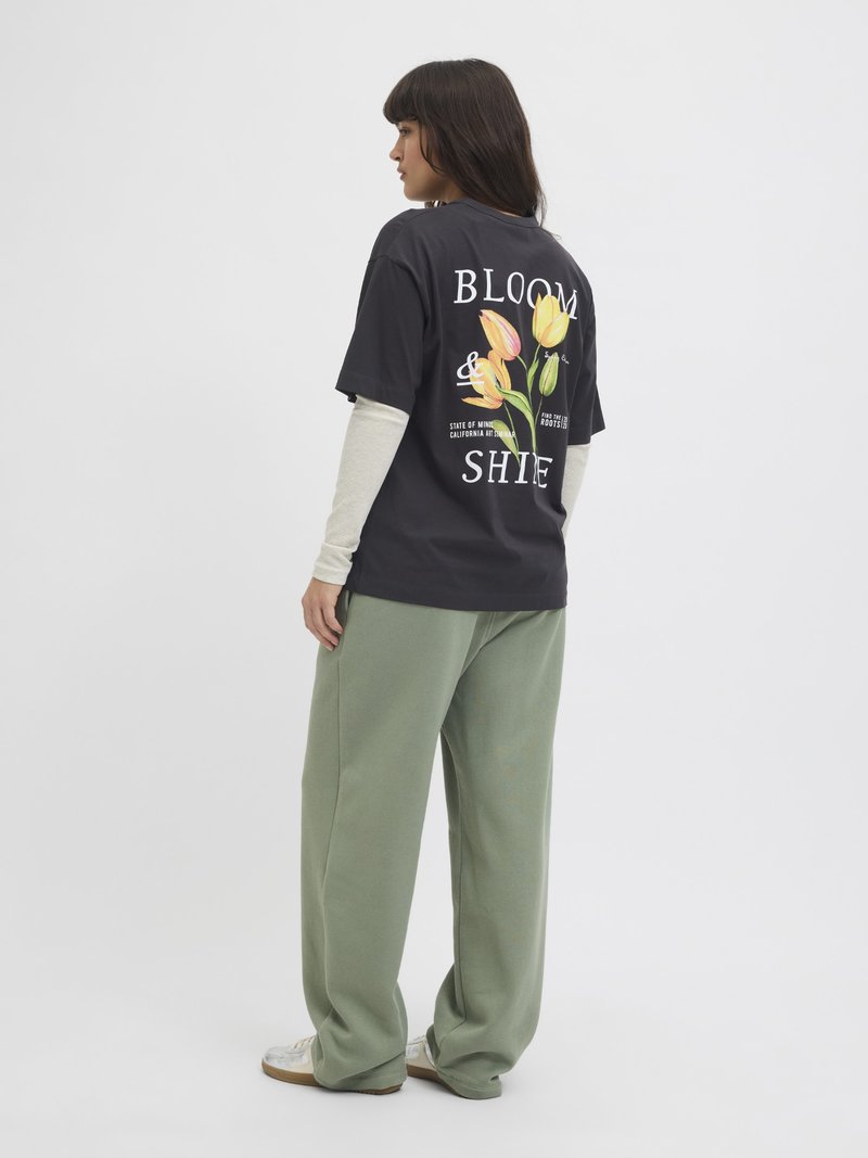 Schwarzes Baumwoll-T-Shirt mit bunten Blumenmotiven und weißem Text auf der Rückseite, kombiniert mit lockeren grünen Sweatpants und weißen Sneakern.