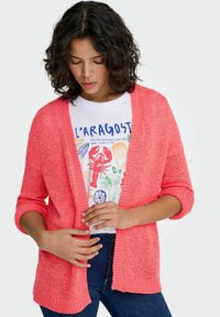 Coral knit cardigan with open front and three-quarter sleeves, featuring a textured pattern and ribbed edges. Worn over a graphic tee.