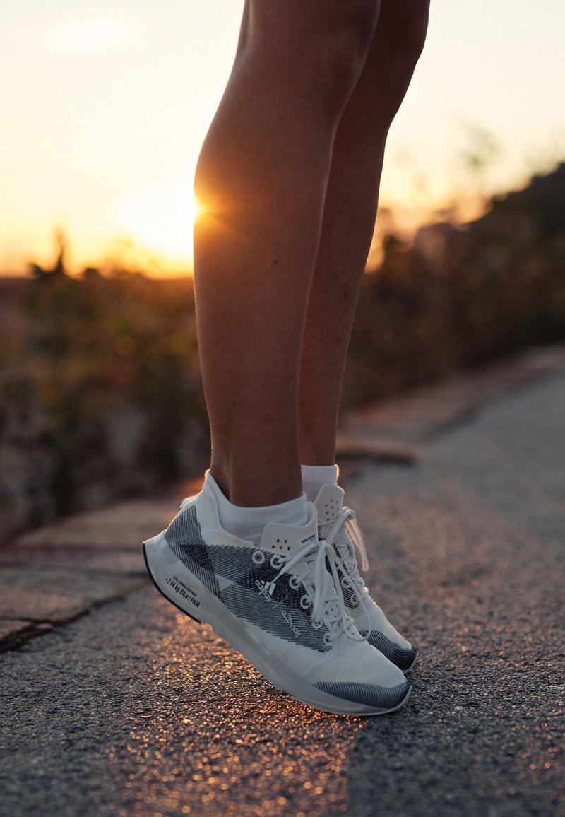 Zapatillas deportivas en blanco y gris con un diseño texturizado, fabricadas con material de malla, cordones planos y suelas de goma, colocadas sobre una superficie pavimentada.