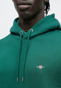 Grøn sweatshirt med hætte, snore med metalender og et lille broderet logo på brystet. Blødt, tekstureret stof.