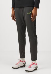 Grijze sportbroeken met een slim fit, Nike-logo in neon groen, gecombineerd met witte en zwarte voetbalschoenen en lichtgekleurde sokken.