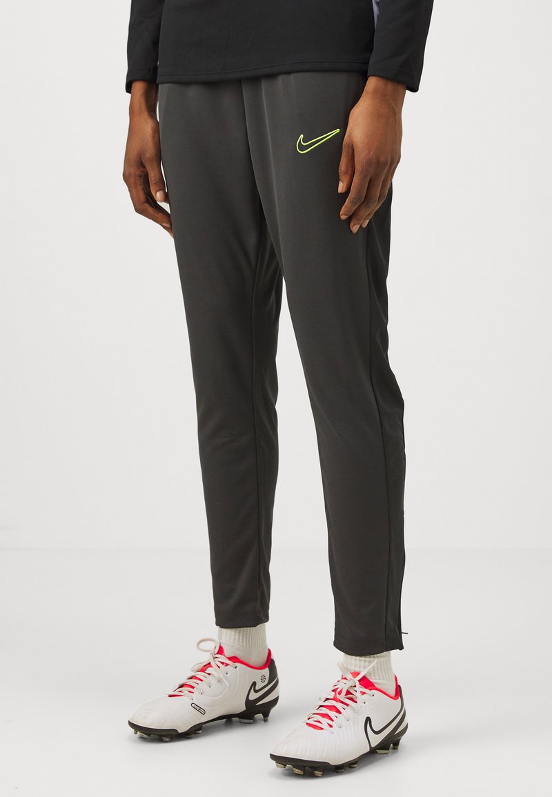 Grijze sportbroeken met een slim fit, Nike-logo in neon groen, gecombineerd met witte en zwarte voetbalschoenen en lichtgekleurde sokken.