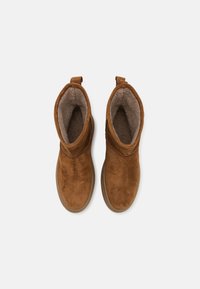 Braune Wildleder-Ankle Boots mit einem weichen grauen Futter, einer glatten, abgerundeten Zehe und einer Zuglasche an der Ferse für ein einfaches Anziehen. Flache Gummisohle.