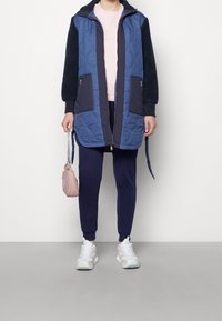 Quilted blå lang jakke med mørkeblå ærmer, parret med pink top, marineblå bukser og hvide sneakers. Har to sidelommer og en lille taske.