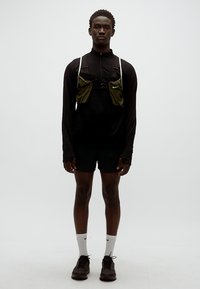 Camisola preta de manga comprida, calções justos, colete de corrida verde, meias brancas e ténis escuros. Design simples, roupa desportiva para desempenho.