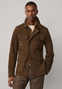 Braune Wildlederjacke mit Kragen, vier vorderen Taschen und Knopfverschluss. Glatte Textur und taillierte Passform, getragen über einem hellen Hemd.