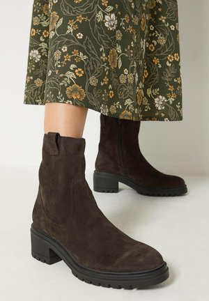 Bottines en daim marron aux semelles épaisses noires portées avec une jupe midi verte à motif floral sur un fond clair uni.
