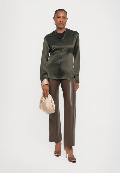 Dark green satin button-up shirt, beige satin clutch, brown leather trousers, and open-toe heels. Smooth textures and tailored fit.