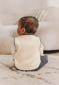 Knit vest in cream with navy accents, worn over a white long-sleeve shirt. Baby seated on a textured light rug with a soft background.