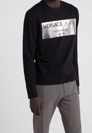 T-shirt noir à manches longues avec un logo métallique argenté dans un encadré portant la mention "Versace Jeans Couture" en police grasse. À assortir avec un pantalon à motif pied-de-poule.