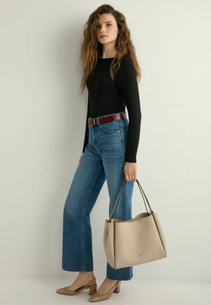 Kobieta z długimi falującymi włosami, ubrana w czarną bluzkę z długim rękawem, niebieskie jeansy, brązowy pasek, beżowe buty na słupku, trzymająca dużą beżową torebkę.