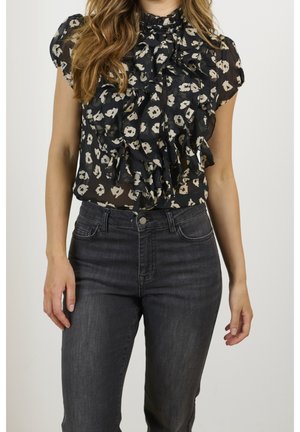 Femme portant un chemisier noir transparent à volants floraux et un jean taille haute gris foncé, debout devant un fond uni.