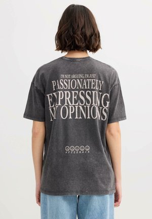 Persona con cabello corto y marrón vista de espaldas, lleva una camiseta negra con la frase "No estoy discutiendo, solo estoy expresando mis opiniones con pasión" con emojis debajo.