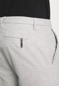 Pantalon gris clair en tissu texturé avec coupe droite. Comprend une poche arrière avec une petite étiquette noire et des accents de couture.