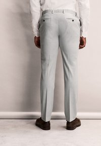 Pantaloni grigi chiari su misura con una texture liscia, due tasche posteriori, dettagli in bottone nero e un design a gamba dritta. Indossati con scarpe marroni.