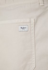 Bež pamukasti materijal s bočnom džepom koji ima plavu i bijelu oznaku s natpisom "Pepe Jeans London." Jednostavno šivanje duž rubova.