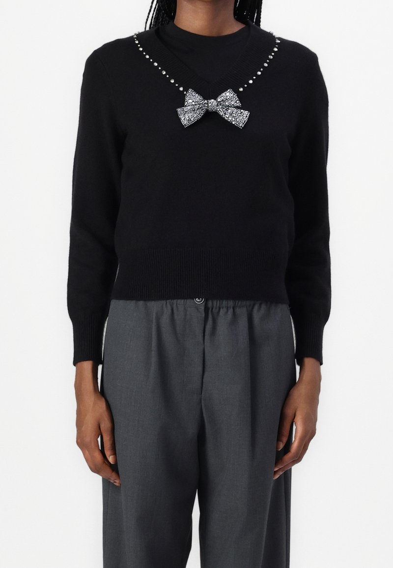 Czarny sweter z dekoltem w serek, ozdobiony srebrnymi ćwiekami i srebrną, cekinową kokardą przy szyi. Noszony z szarymi, luźnymi spodniami.