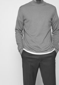 Uomo che indossa un pullover a maniche lunghe con colletto tondo sopra una maglietta bianca visibile, con le mani nelle tasche di pantaloni su misura.