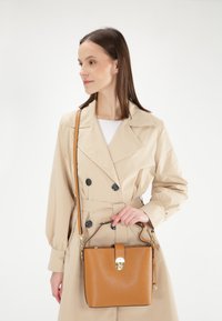Tan læderhåndtaske med en struktureret form, guldfarvede detaljer og en aftagelig rem, præsenteret sammen med en beige trenchcoat.