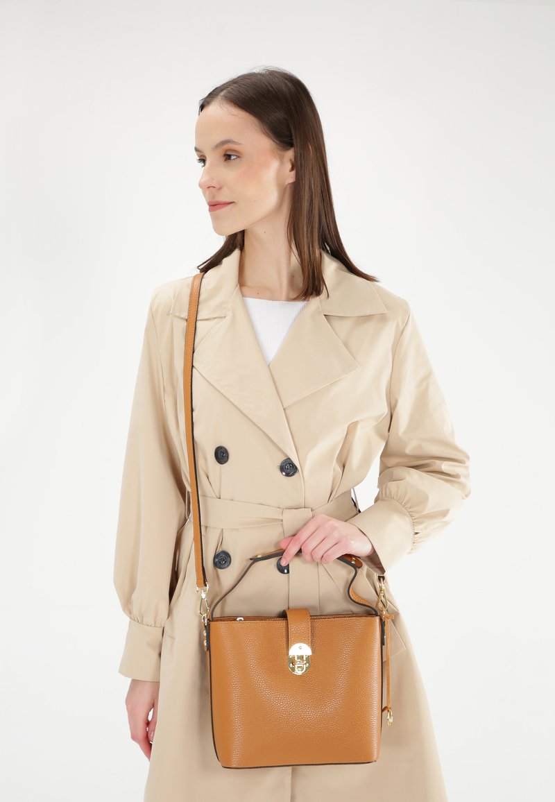Tan læderhåndtaske med en struktureret form, guldfarvede detaljer og en aftagelig rem, præsenteret sammen med en beige trenchcoat.