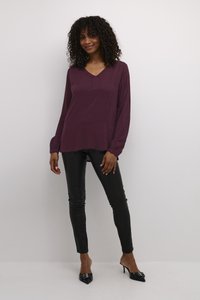 Langärmlige, pflaumenfarbene Bluse mit V-Ausschnitt und subtilen Falten-Details, kombiniert mit engen schwarzen Lederhosen und spitzen Pumps.