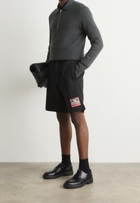 Pull zippé en charbon associé à un short noir avec un logo HUGO rouge et blanc. Chaussures en cuir noir complètent la tenue.