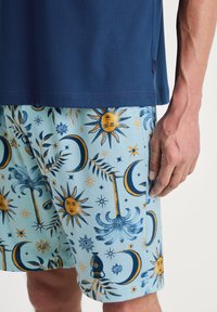 Korte mouwen marineblauw shirt onder een lichtblauwe broek met een patroon van zon, maan en palmmotieven in geel en donkerblauw. Van katoen materiaal.