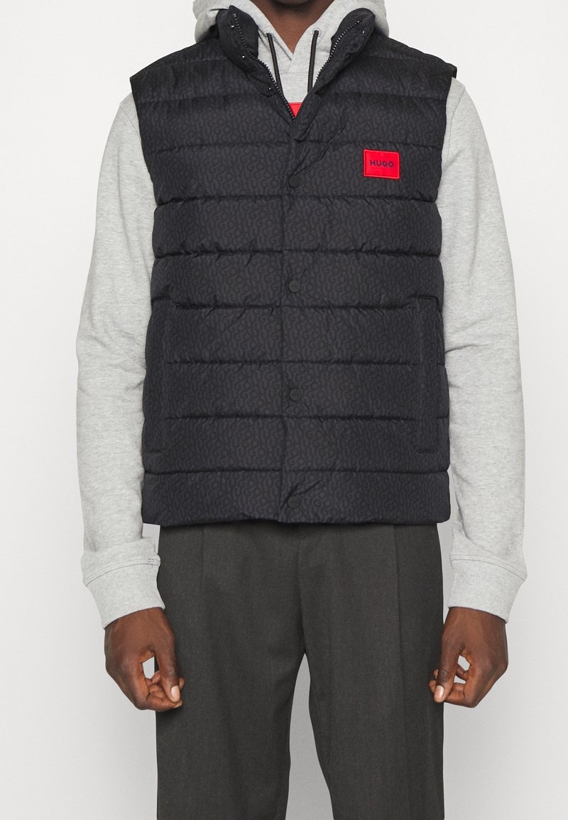 Schwarze gepolsterte Weste mit einem dezenten Muster, ausgestattet mit einem roten Logo-Patch, Knöpfen auf der Vorderseite und einer Kapuze. Über einem grauen Hoodie getragen.