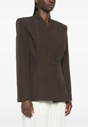 Vrouw draagt een gestructureerde donkerbruine blazer met gewatteerde schouders en een enkele knoop, gecombineerd met witte geplooide broek.