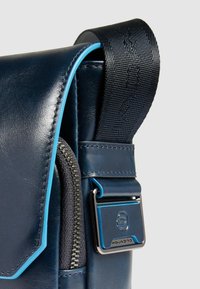 Marineblaue Ledertasche mit hellblauem Akzent auf der Klappe und einem strukturierten Stoffriemen mit silberner Schnalle und Reißverschlussdetail.