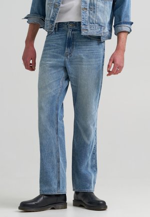 Homme portant un jean denim bleu clair, une veste en denim assortie avec les manches retroussées, une chemise blanche et des bottes en cuir noir, debout.
