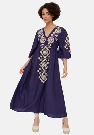 Frau trägt ein bodenlanges, marineblaues Kleid mit cremefarbenen Blumenstickereien und gewellten Kanten, kombiniert mit beigen Keilschuhen, steht vor weißem Hintergrund.